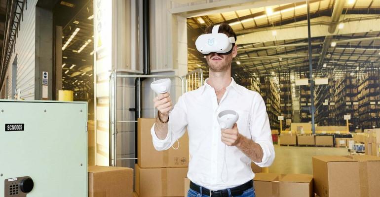 A man in a white shirt wearing a VR headset with VR controllers. A warehouse interior filled with cardboard boxes in the background.