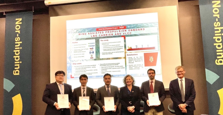 Representatives of the JDP companies stand with their AiP certificates in front of a projected power point slide on a stage with Nor Shipping branding