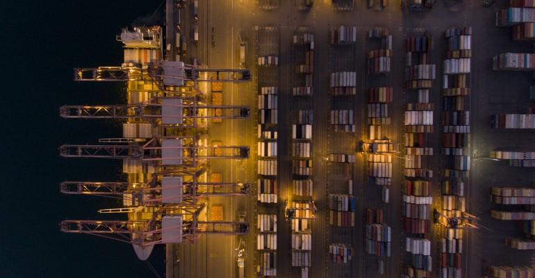 An overhead shot of a container ship at berth at night, gantry cranes and container stacks.