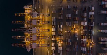 An overhead shot of a container ship at berth at night, gantry cranes and container stacks.